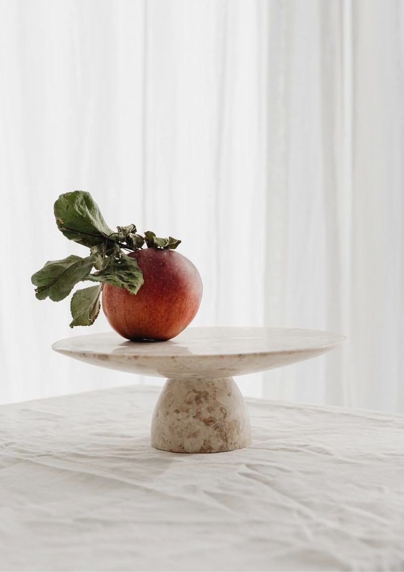 A red apple still on its leafy stem, resting on a stone cake stand on white linen