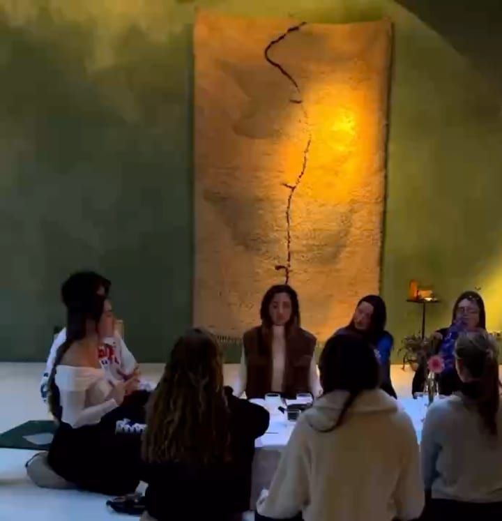 Women seated on cushions around a long low table lit by candles, one of them speaking